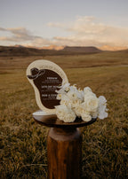 AALVO custom event signage. Decorative cowboy themed bar menu sign with flowers on a wooden stand against a scenic mountainous landscape.