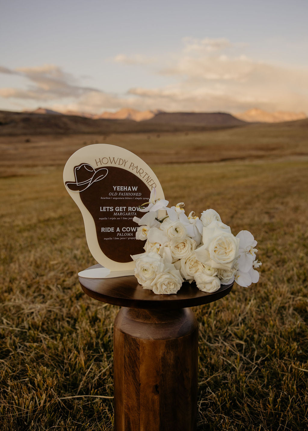AALVO custom event signage. Decorative cowboy themed bar menu sign with flowers on a wooden stand against a scenic mountainous landscape.