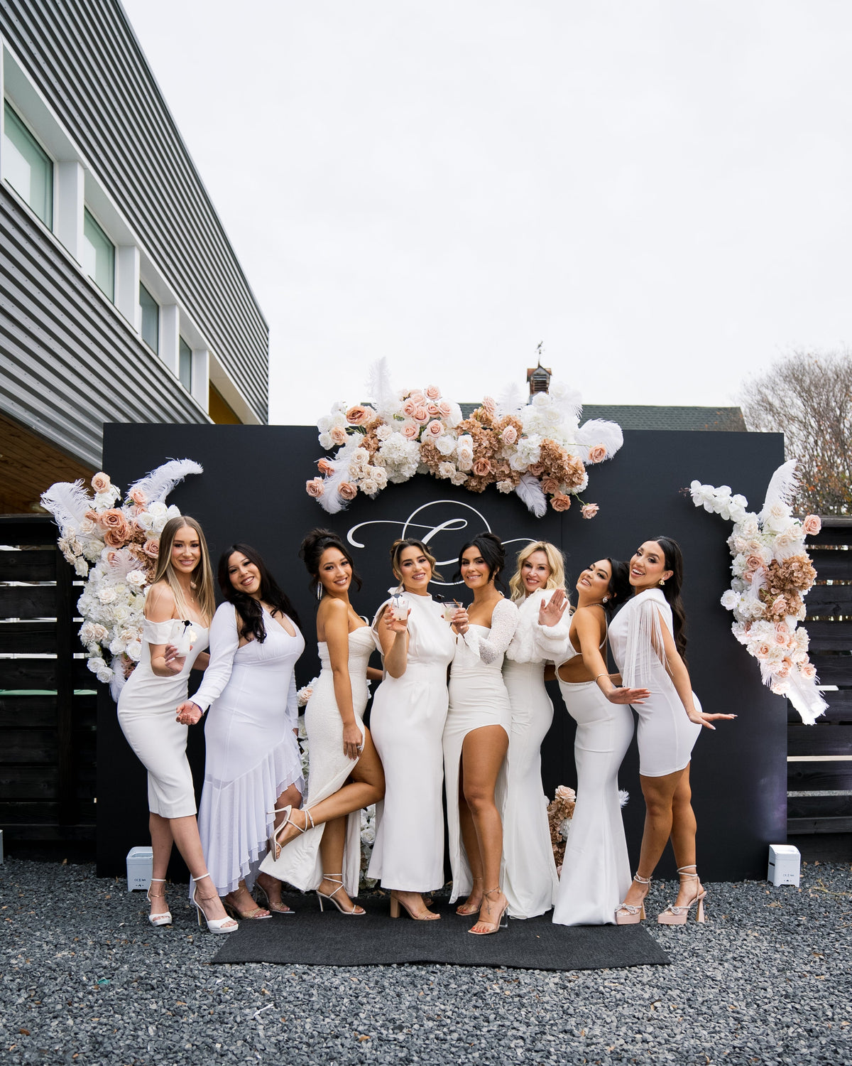 AALVO custom event signage. A group of eight women posing in elegant white and champagne wedding dresses in front of a black decorative sign decorated with large floral arrangements all around the sign.