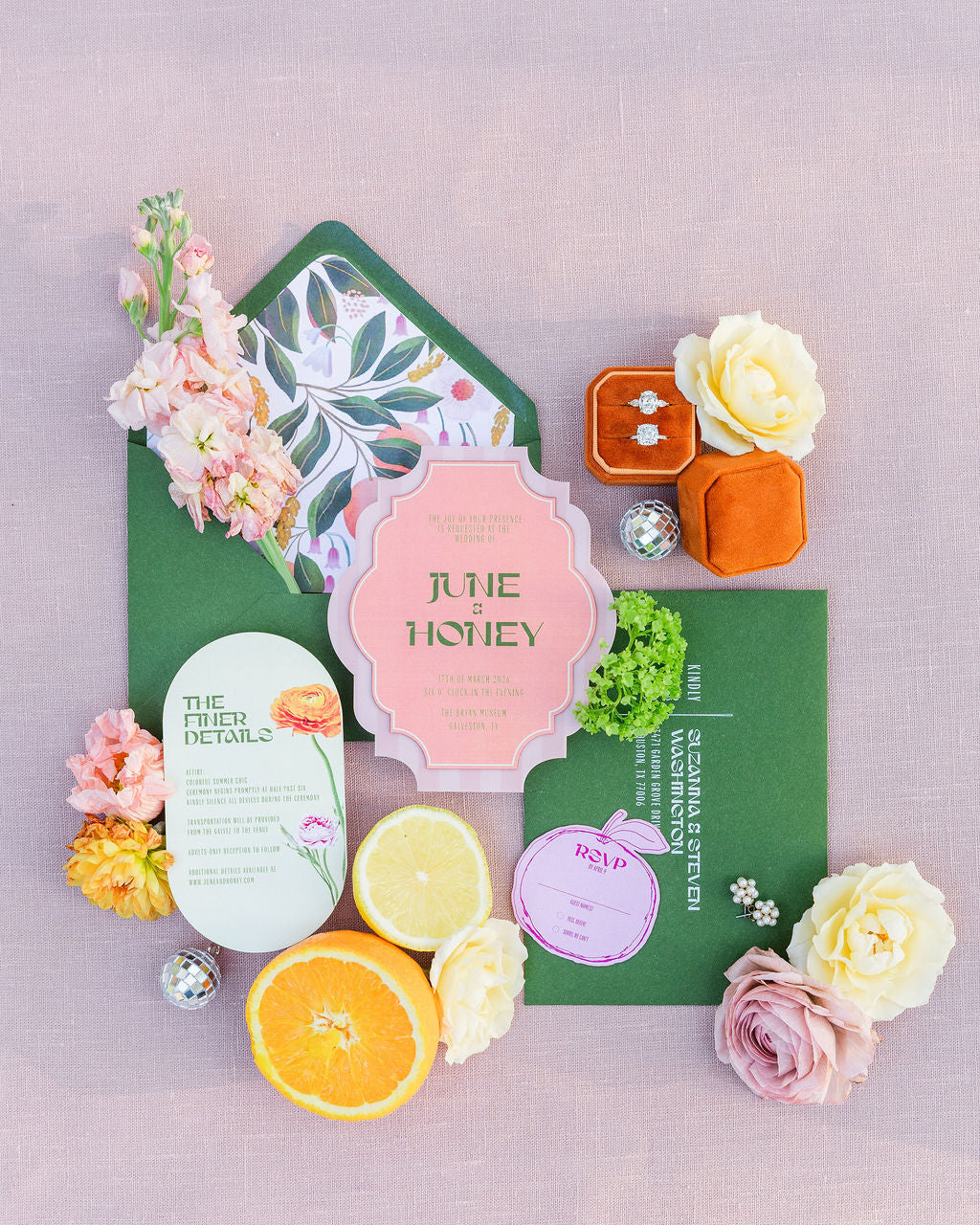 AALVO custom wedding invitation. A styled flat lay of a wedding invitation suite on a dusty pink background, featuring a pink central invitation card, a green envelope with botanical leaf patterns, a green details card, a purple RSVP card, floral and citrus garnishes, and a small orange velvet ring box with diamond rings inside.