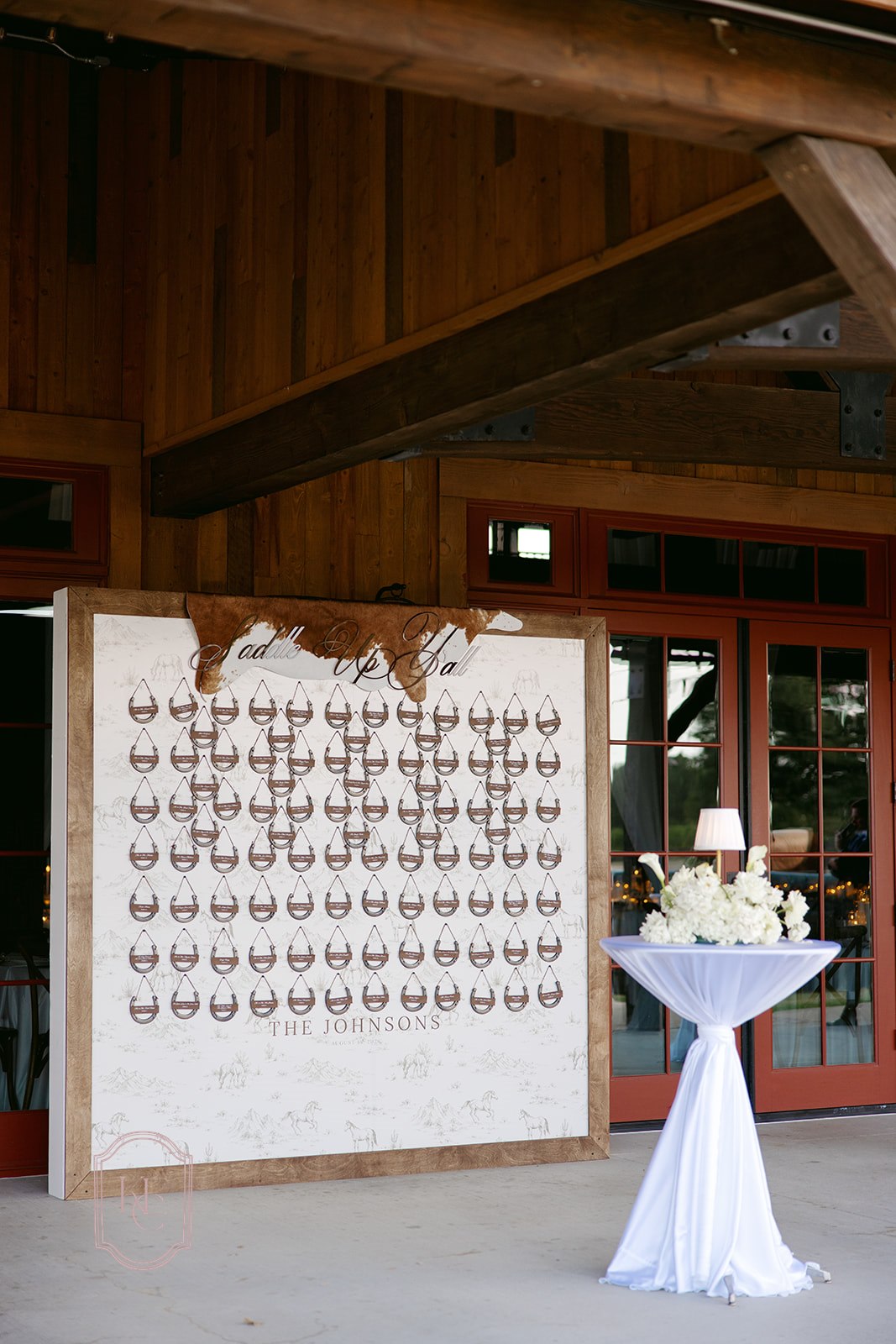 AALVO custom wedding signage. A rustic wedding reception seating chart backdrop mounted on a wooden wall, featuring rows of horseshoes with names on them. A draped white table with floral arrangements stands beside it in front of red-framed glass doors.