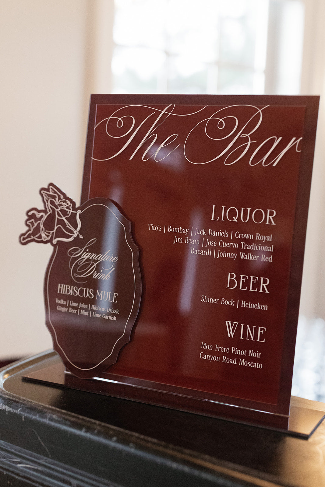 AALVO custom wedding signage. A custom acrylic bar menu sign for 'The Bar' featuring a dark reddish-brown background with white script and text listing liquor, beer, and wine selections.
