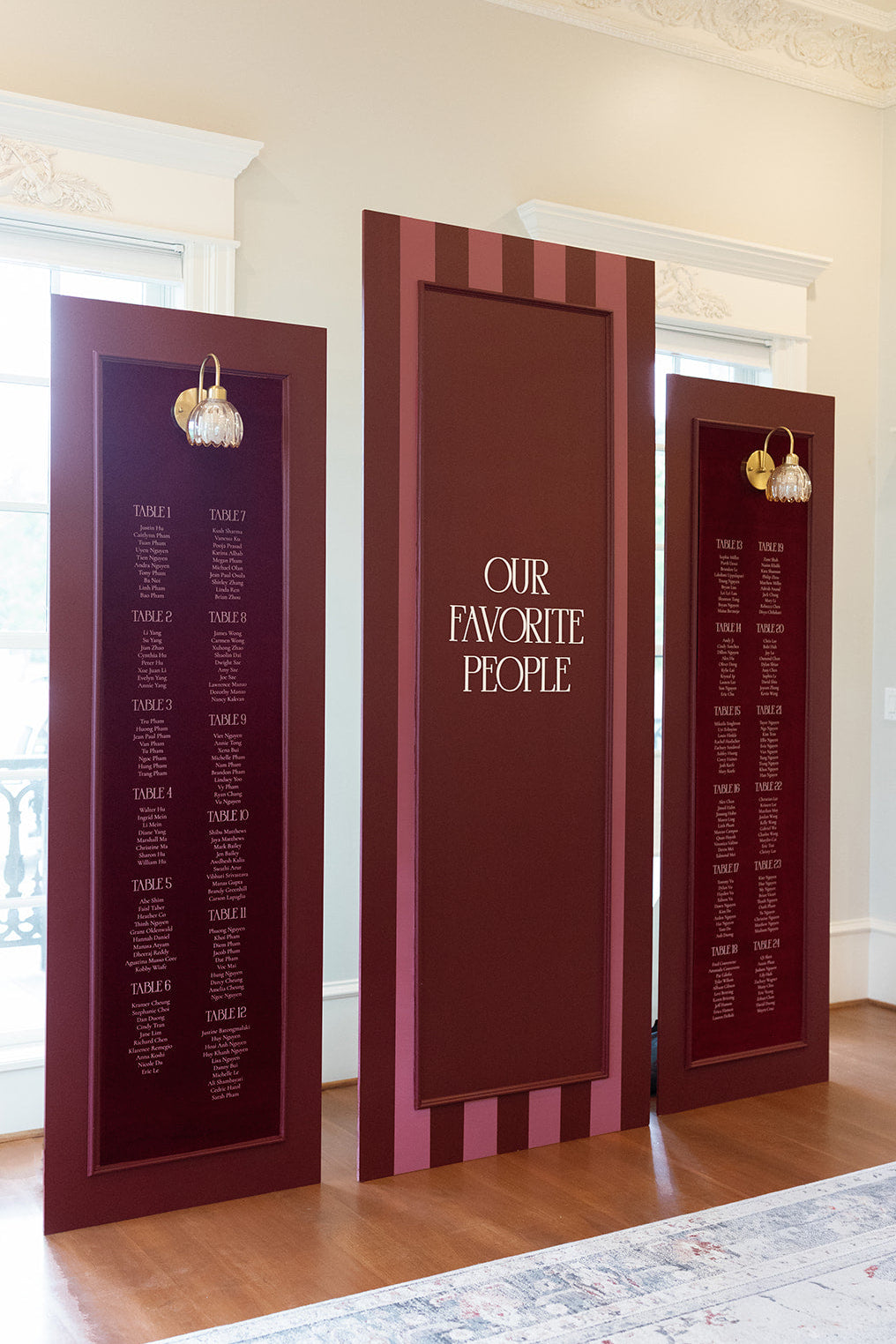 AALVO custom wedding signage. Three freestanding maroon wedding guest seating chart panel signs with white text, displayed in a bright room with wood floors and cream walls.