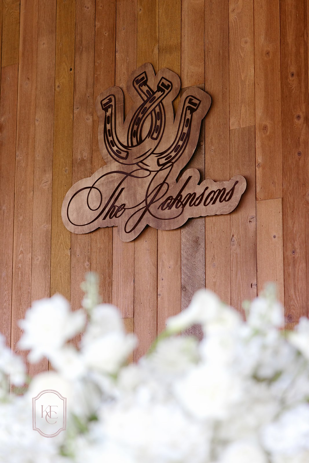 AALVO custom wedding signage. A custom laser-cut wooden sign featuring two horseshoes above the script name 'The Johnsons', mounted on a vertical wood plank wall with blurred white flowers in the foreground.