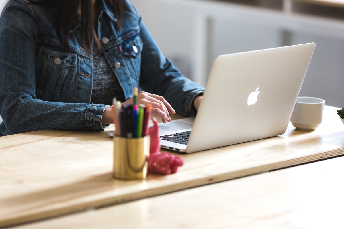 Woman sitting at a desk working on a macbook laptop, with coffee mug and pens near her.